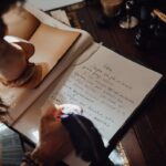 From above of crop unrecognizable female writer taking notes in copybook with feather at vintage table in sunbeam