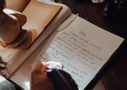 From above of crop unrecognizable female writer taking notes in copybook with feather at vintage table in sunbeam