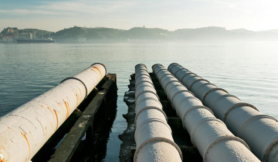 Sewage pipes stretch over a tranquil waterfront in Lisbon, highlighting urban infrastructure.
