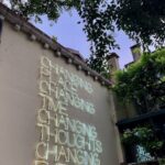 Neon sign with motivational text on a building in Italy surrounded by greenery.
