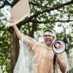 Man holding megaphone and cardboard at protest rally in park.